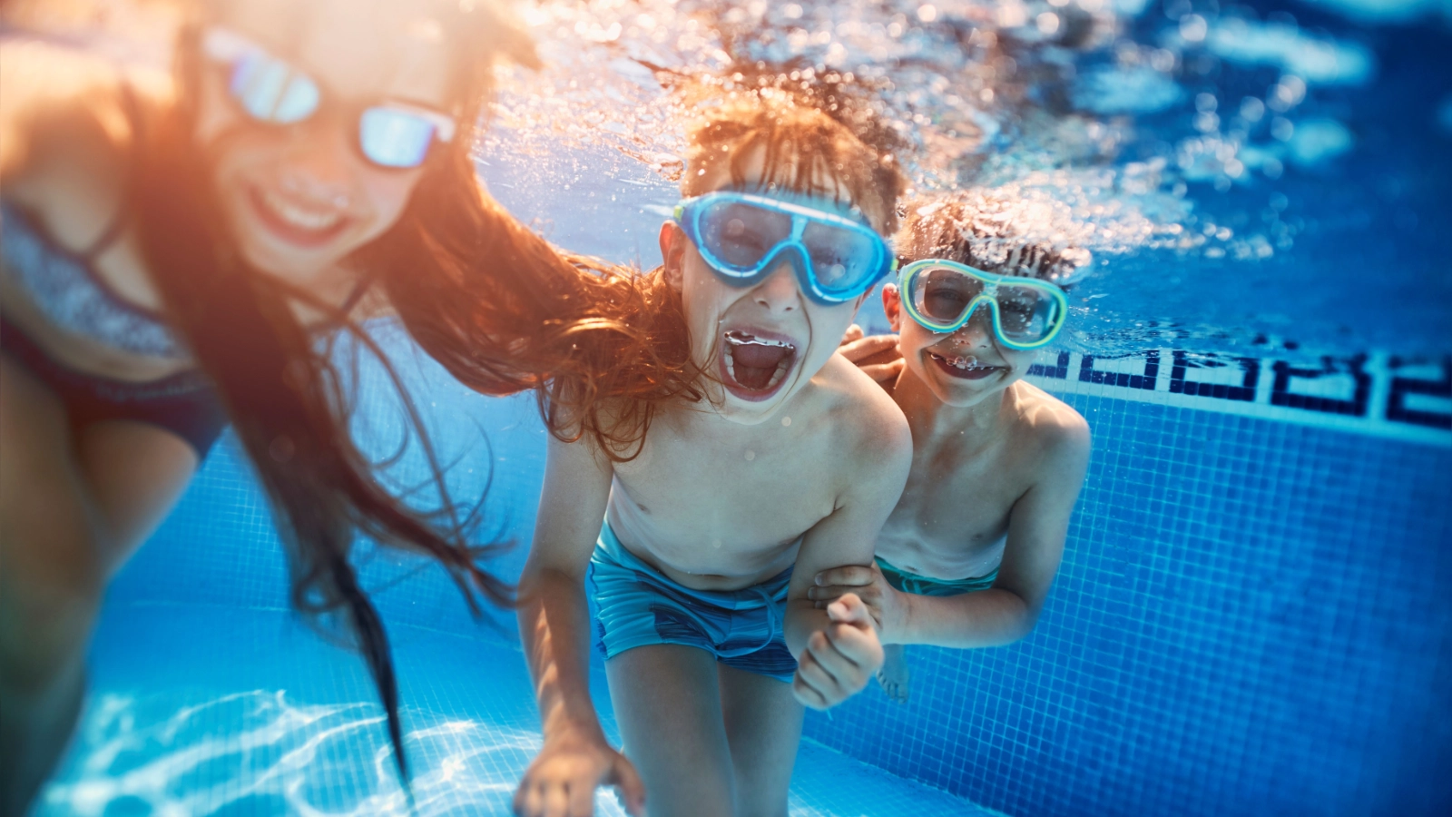 Three kids swimming underwater in a pool wearing goggles.